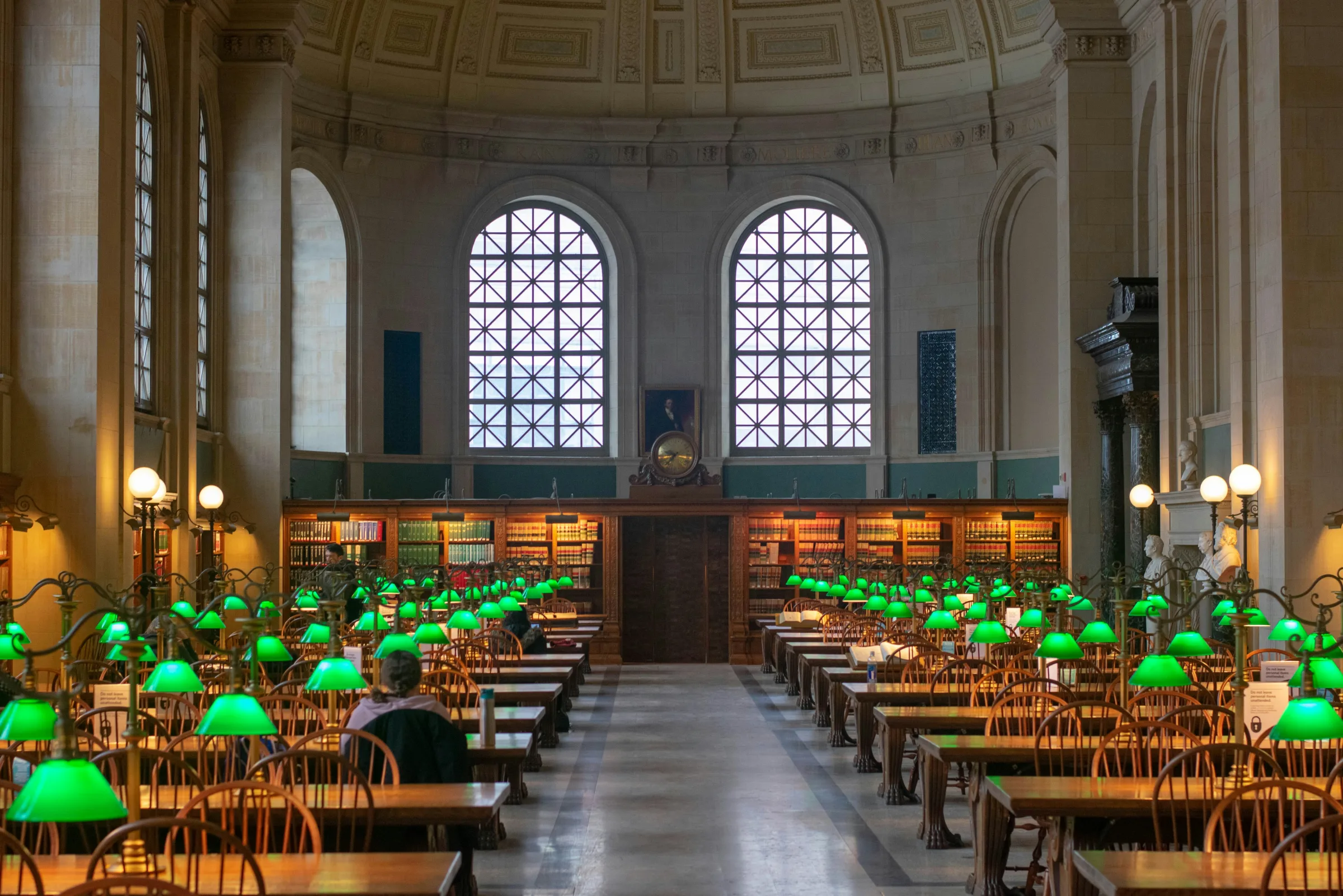 An ornate library reading room with rows of green lamps and bookshelves.